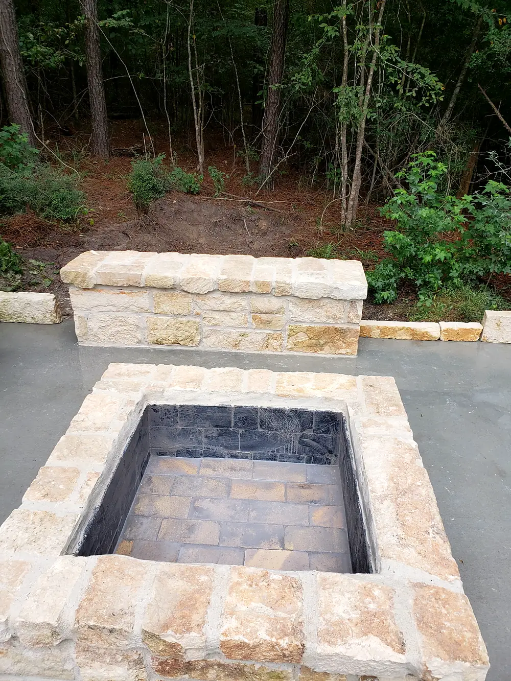 Square chopped-limestone fire pit with a matching stone bench on a new slab, woods behind
